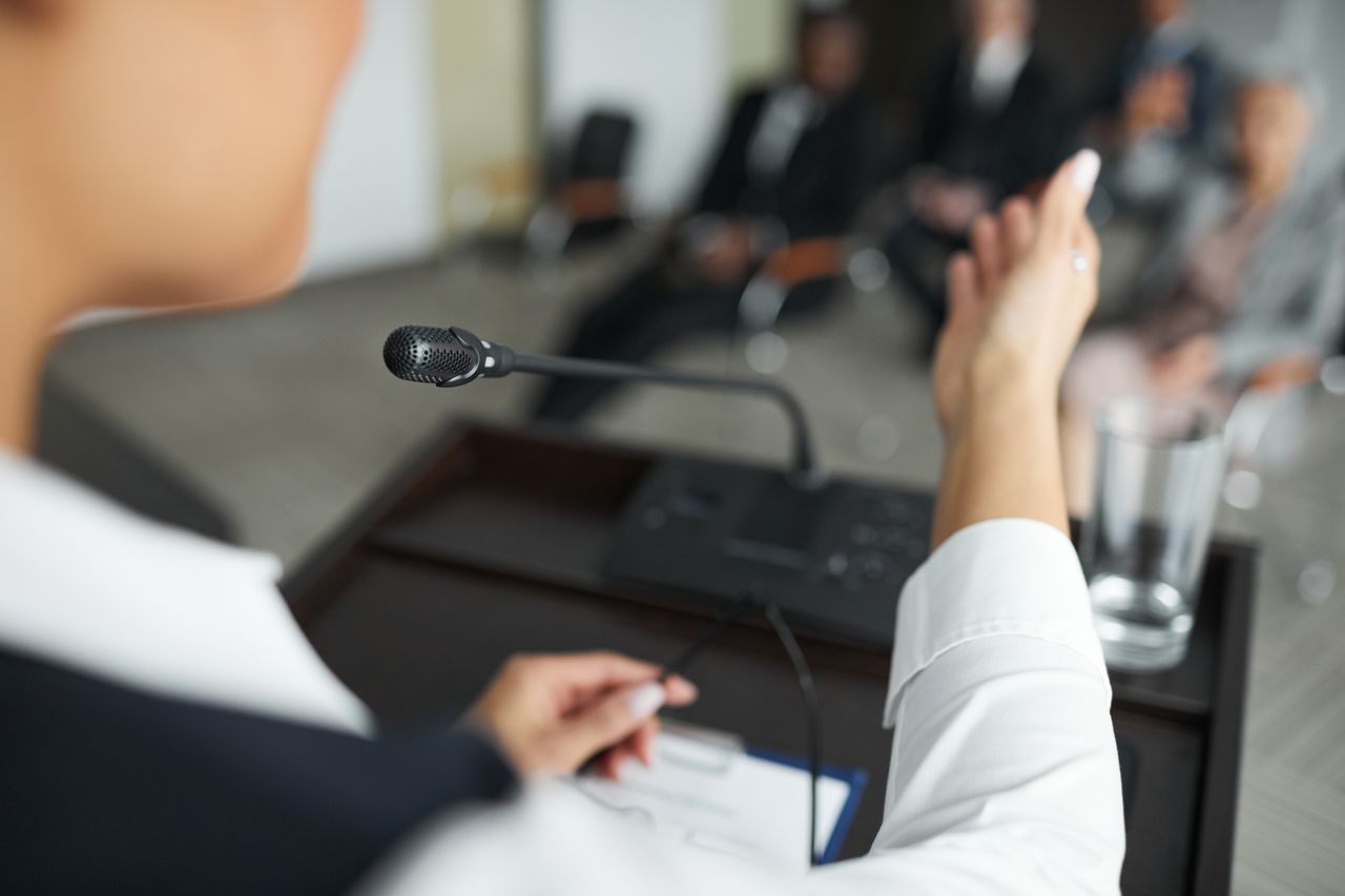 Service A person speaking at a podium with a microphone to an audience during a business meeting.